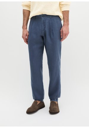 Mężczyzna ubrany w niebieskie spodnie na miarę, beżowy sweter z dzianiny i brązowe zamszowe buty wsuwane, stojący na jednolitym jasnym tle.