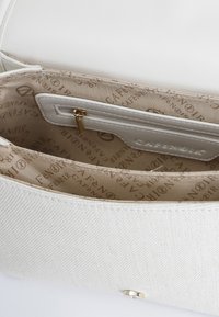 Vit handväska med texturerat yttermaterial. Inredningen har en beige foder med mönster av 'Café Noir'-logotyper och en liten dragkedjeficka.