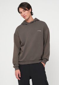 Barna kapucnis pulóver vastag anyagból, bordás mandzsettákkal és szegéllyel, apró logóval a mellkasán. Fekete nadrággal viselve.