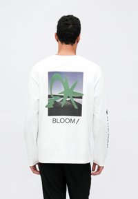 Weißes langärmeliges T-Shirt mit grafischem Druck in grünen Formen und dem Wort "BLOOM/" auf der Rückseite. Glatter Stoff, lockerer Schnitt.