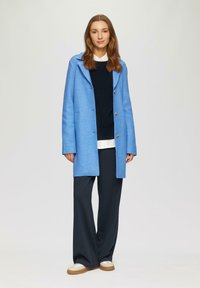 Cappotto di lana blu chiaro con cappuccio, grandi bottoni e taglio morbido. Abbinato a un maglione blu navy, pantaloni a gamba larga e scarpe bianche.
