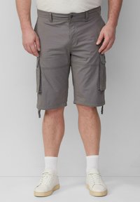 Mann in grauen Cargo-Shorts mit Seitentaschen, weißen Socken und weißen Turnschuhen, der vor einem schlichten grauen Hintergrund steht.