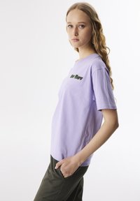 Lila katoenen T-shirt met korte mouwen en een ronde hals, voorzien van groene geborduurde tekst "Go Flow" op de voorkant. Model draagt een donker groene broek.