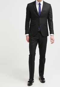 Completo nero con una vestibilità su misura, caratterizzato da una giacca monopetto, rever a golfo e pantaloni slim. Abbinato a una camicia bianca e una cravatta blu.