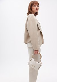 En kvinna står i profil, iförd en beige oversize-jacka och krämfärgade byxor, medan hon håller en vit handväska med flätat handtag.