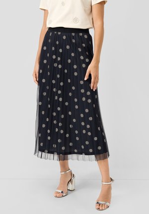 Kobieta w czarnej plisowanej spódnicy midi w szare groszki, beżowym topie z drobnymi kwiatowymi wzorami oraz srebrnych sandałach na wysokim obcasie.