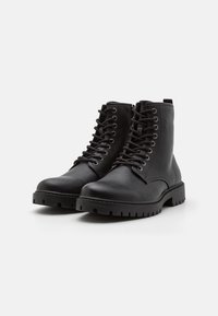 Botas de tobillo de cuero negro con puntera redonda, suela de goma plana, ojales metálicos y cierre de cordones en la parte delantera. Textura suave con un acabado mate.