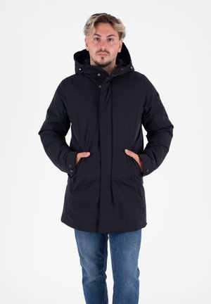 Zwarte, hooded jas met knoopsluiting en zijzakken; gemaakt van een gladde, waterafstotende stof. Draagt over jeans.