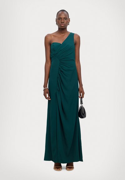 Robe maxi teal à une épaule avec un tissu drapé et des détails de fronces. Le mannequin tient un petit sac à main noir, portant des accessoires minimalistes.
