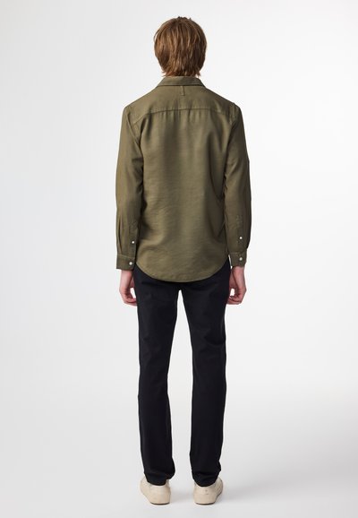 Olivengrøn skjorte med lange ærmer og knapmanchetter, buet søm og en glat tekstur, parret med sorte bukser og lysfarvede sko.