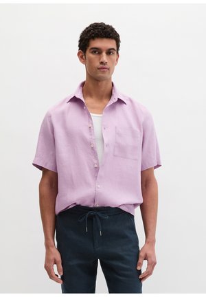 Junger Mann, der ein hellviolettes kurzärmeliges Hemd über einem weißen Tanktop und dunkle Hose mit Kordelzug vor einem schlichten Hintergrund trägt.