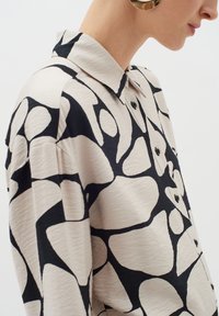 Kerge kangaäärisega nööbiga särk, millel on beež taust ja julge must abstraktne muster, koos krae ja nööbiga detailidega.