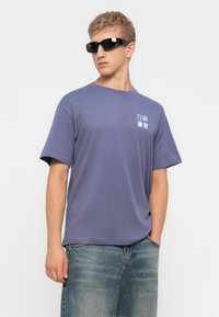 Joven con gafas de sol negras envolventes, camiseta gráfica morada con pequeño texto blanco y rojo, y jeans azules desgastados, de pie con una mano en el bolsillo.