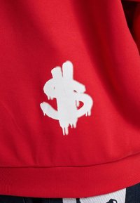 Indumento rosso con un grande logo di dollaro sprayato in bianco con gocce. Texture liscia, orlo inferiore a coste.