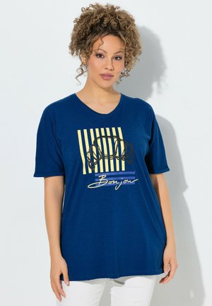 Donna con capelli ricci indossa una T-shirt blu con strisce gialle, una grafica nera di croissant e la parola "Bonjour" in corsivo.