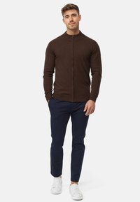 Brauner Reißverschluss-Pullover in enganliegendem Design, kombiniert mit marineblauen Hosen. Weiße Sneaker vervollständigen das Outfit. Glatte Textur, lässiger Stil.