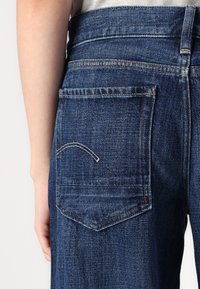 Dark blue denim jeans feature a back pocket with contrasting stitching and a curved design. The fabric has a textured, woven appearance.