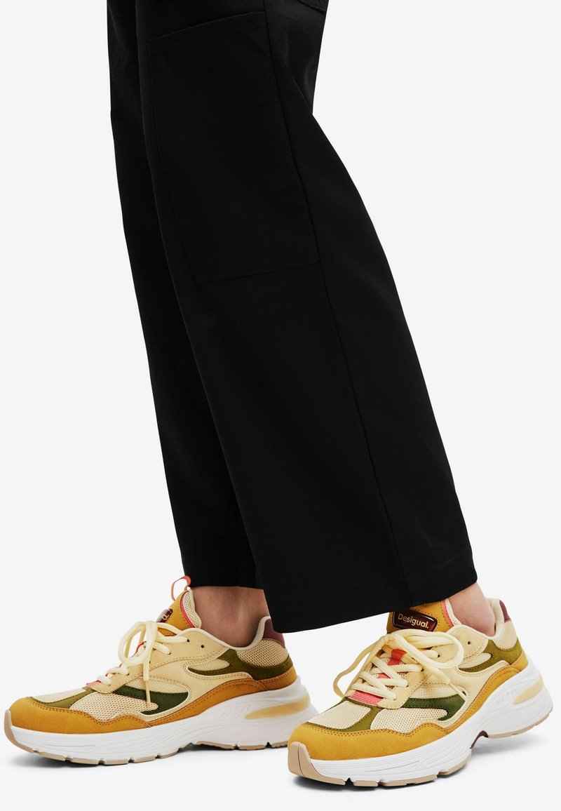 Pieds chaussés de baskets beige, moutarde et vert olive avec des semelles blanches, assortis à un pantalon noir à jambes larges sur fond blanc.