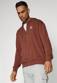 Brązowy hoodie na zamek z miękkiego materiału, z kieszenią kangurzą, prążkowanymi mankietami i haftowanym logo na piersi.