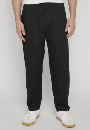 Uomo che indossa pantaloni neri su misura e sneaker bianche, in piedi su uno sfondo chiaro e semplice, mani rilassate ai lati.