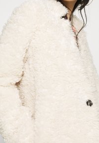 Hvid jakke i imiteret pels med en tekstureret overflade, der har en rundet krave og en enkelt sort knapdetalje. Enkel, firkantet silhuet.