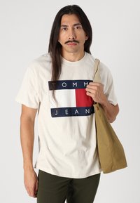 Kreemivärviline puuvillane t-särk, millel on sinine ja punane logo, millel on tekst "TOMMY JEANS". Modell hoiab käes khaki värvi kotti.