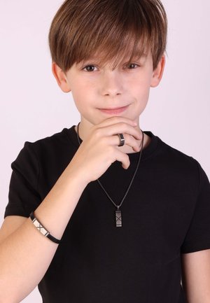 Zwarte T-shirt model dat een zwarte metalen hangerketting en een bijpassende geometrische ring en armband draagt, met textuurelementen.