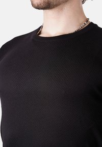 Suéter negro de textura con cuello redondo, que presenta un patrón de panal. Un collar con cuentas de plata es parcialmente visible.