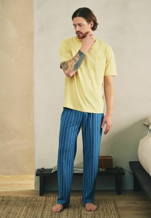 Man with tattooed arm wearing yellow t-shirt and blue striped pants standing barefoot on rug in minimalist bedroom.