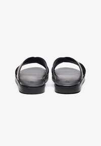 Une paire de sandales noires en cuir à glissière avec de larges brides croisées et des semelles plates, vues de face sur fond blanc.