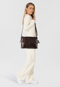 Bolso mensajero de cuero marrón oscuro con acentos de doble hebilla, asa superior, correa ajustable y dos bolsillos frontales. Textura suave, forma rectangular.