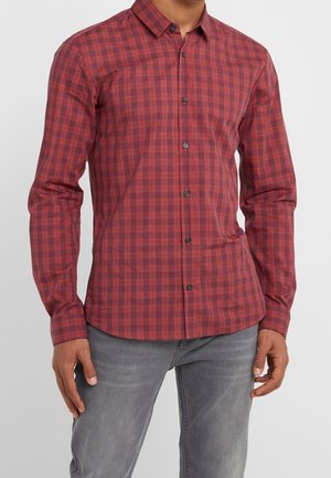 Mand iført en tætsiddende langærmet rød- og sortternet skjorte med knapper og grå jeans, stående mod en ensfarvet baggrund.