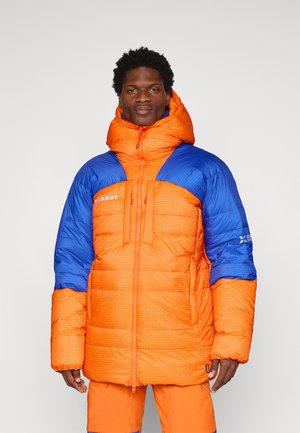Casaco acolchoado com cores laranja e azul brilhantes, apresentando um padrão de grelha, gola alta e fecho com zip. Inclui um logótipo e grandes bolsos.
