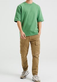 Zelené bavlněné tričko s krátkým rukávem a kulatým výstřihem, spárované s béžovými cargo kalhotami s několika kapsami a šedými sportovními botami.