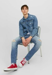 Chaqueta de denim con ligeras desgastadas, combinada con jeans rotos de color azul claro. Zapatillas rojas con suelas blancas. El modelo está sentado en una silla metálica.