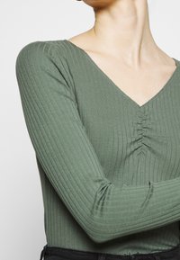 Top à manches longues côtelé vert olive avec un col en V et un détail froncé au niveau du bust, doté d'un tissu doux et texturé et d'une silhouette ajustée.