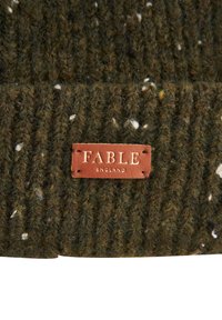 Adīta cepure tumši zaļā krāsā ar baltiem plankumiem, kurai ir brūna ādas etiķete ar iespiestu uzrakstu "FABLE ENGLAND". Teksturēts rievots dizains.