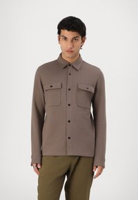 Camisa castanha de mangas longas com colarinho abotoado, dois bolsos no peito e botões pretos. Textura suave com um corte ajustado.
