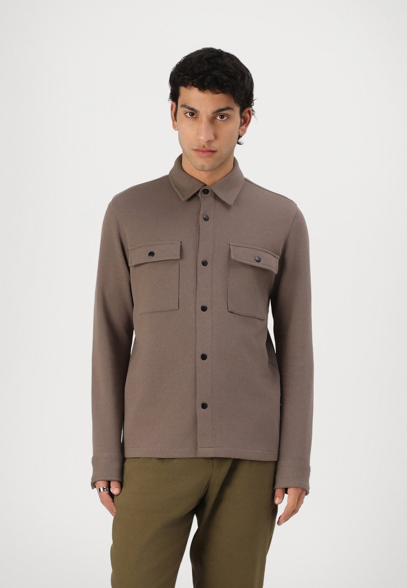 Camisa castanha de mangas longas com colarinho abotoado, dois bolsos no peito e botões pretos. Textura suave com um corte ajustado.