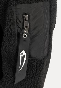 Chaqueta de forro polar negra con un bolsillo lateral con cremallera, fabricada en tejido sintético negro y una etiqueta tejida con un logo blanco. Apariencia texturizada y cálida.
