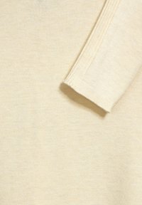 Jasno-beżowa dzianina o gładkiej teksturze, z ryflowanym wykończeniem rękawów. Cały projekt jest prosty i bezszwowy.