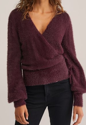 Femme portant un pull cache-cœur duveteux de couleur violet foncé avec de longues manches ballon et des poignets ajustés, assorti à un pantalon noir.