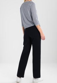 Kvinna i svarta vida byxor, en vit och marinrandig t-shirt med trekvartslång ärm och vita sneakers, står vänd bort.