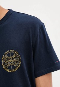 Camiseta de algodón azul marino con mangas cortas, que presenta un logo dorado "TOMMY JEANS NEW YORK CITY" y un pequeño acento tricolor en la manga.