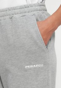 Ranka kišenėje šviesių pilkų sportinių kelnių su elastine juosta ir baltu siuvinėtų "PEGADOR" logotipu ant šlaunies.