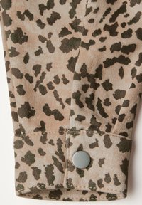 Tissu imprimé léopard brun et vert avec une texture lisse, comportant un poignet avec un bouton-pression argenté et des détails de couture visibles.