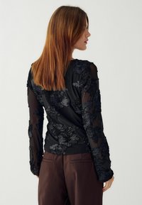 Vrouw met steil bruin haar, gekleed in een doorzichtig zwart longsleeve shirt met 3D-bloemenapplicaties en donkerbruine broek, die zijwaarts staat.