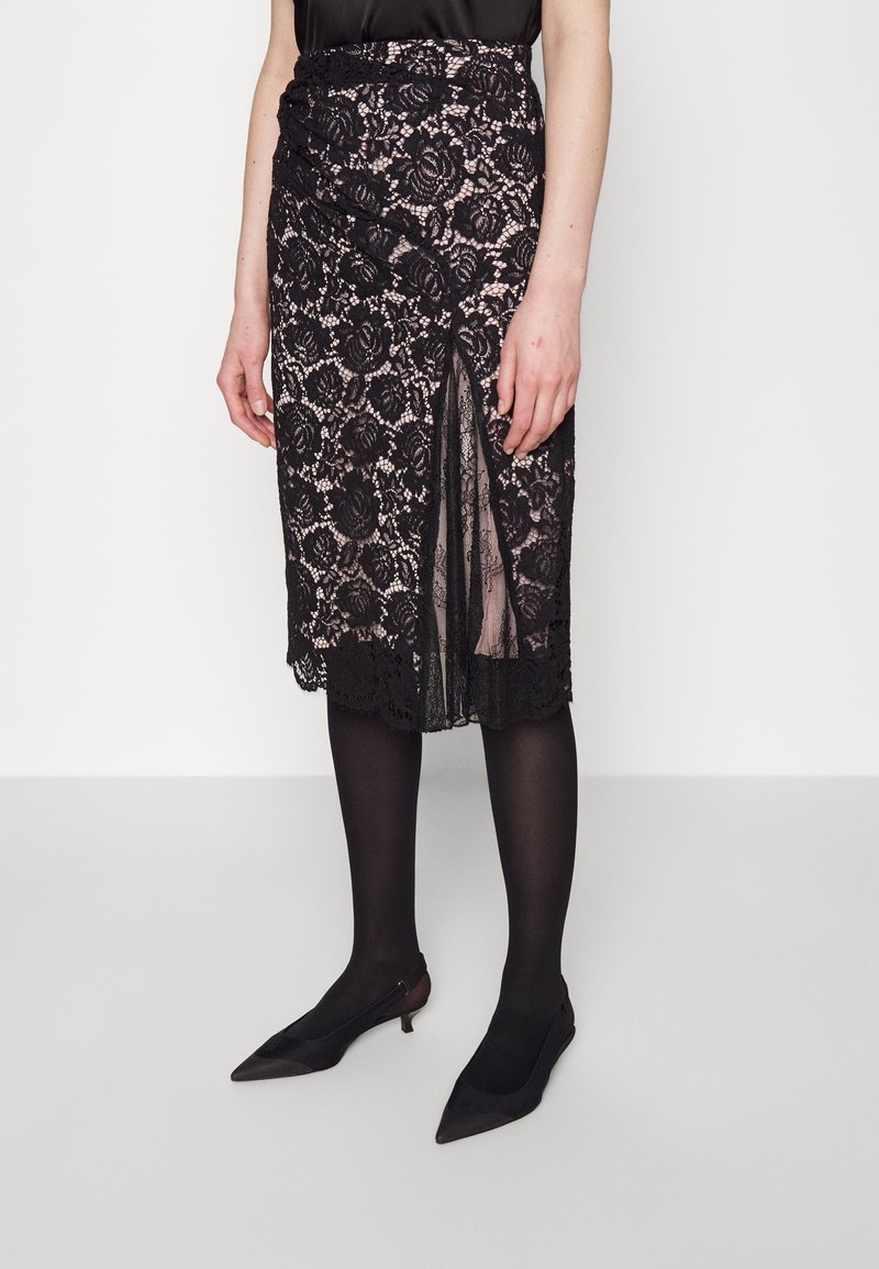 Jupe en dentelle noire longueur genou avec motif floral et fente sur le devant, portée avec des collants noirs et des chaussures noires à talons bas pointues.