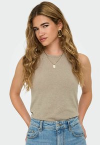 Top sans manches beige avec une finition texturée, associé à un jean taille haute bleu et des colliers en or superposés. Des boucles d'oreilles audacieuses ajoutent une touche de détails.
