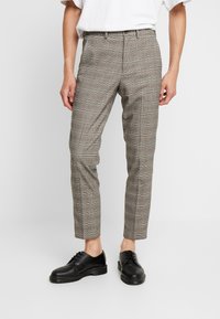 Man i slim-fit grå rutiga byxor, kombinerat med svarta snörskor och en vit kortärmad skjorta, som står mot en neutral bakgrund.